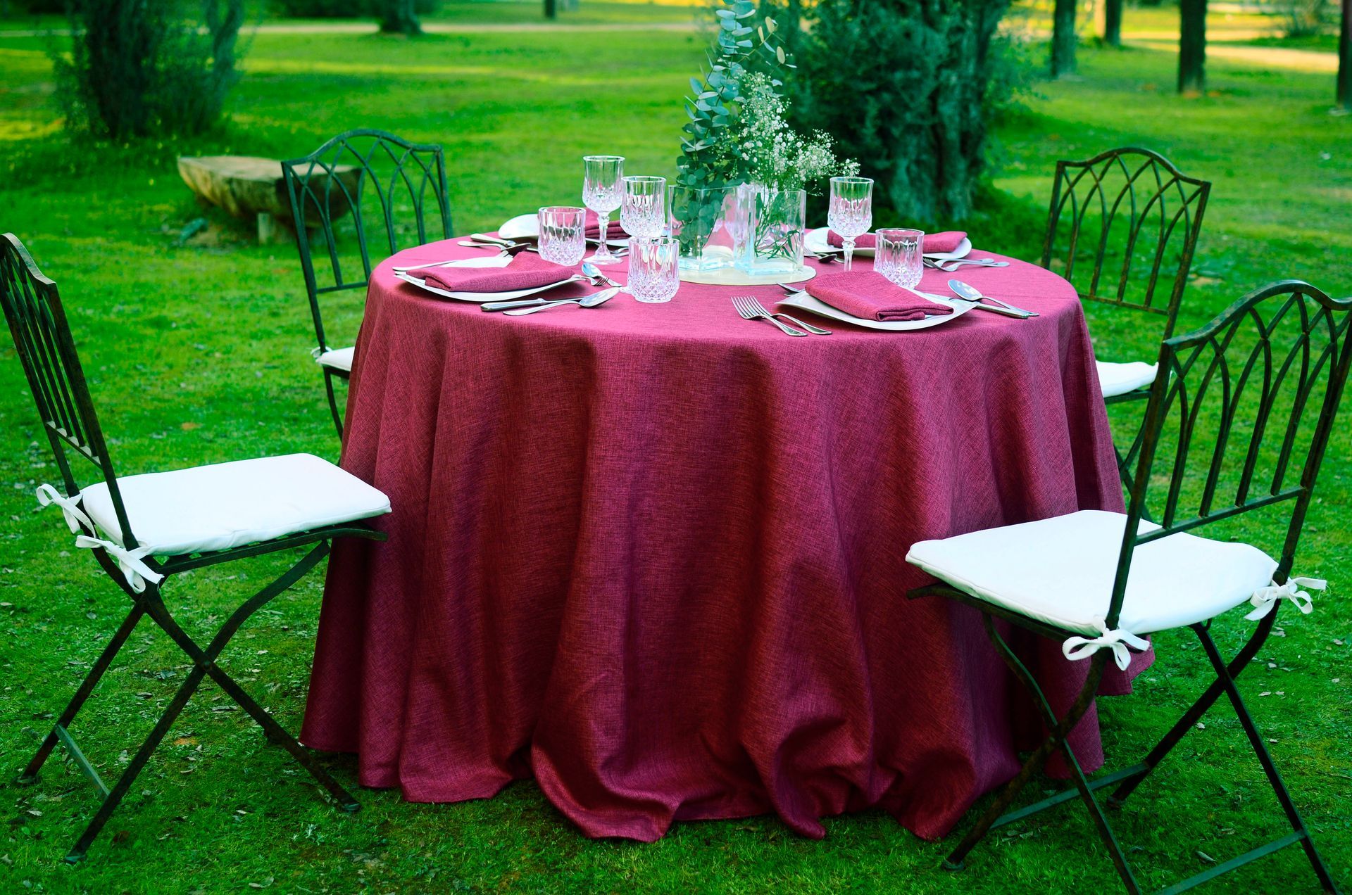 Mesa redonda dispuesta para comer al aire libre, sobre césped, con mantel burdeos y sillas negras.