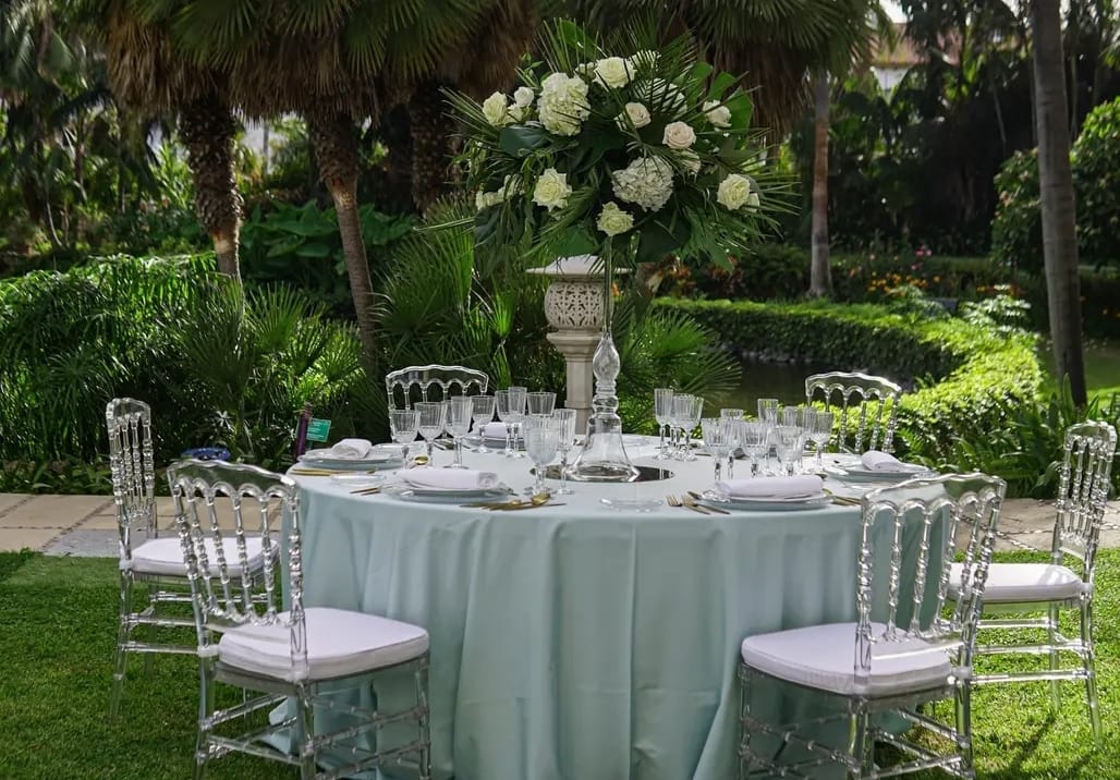 Una mesa redonda con un mantel azul y sillas transparentes en un jardín.