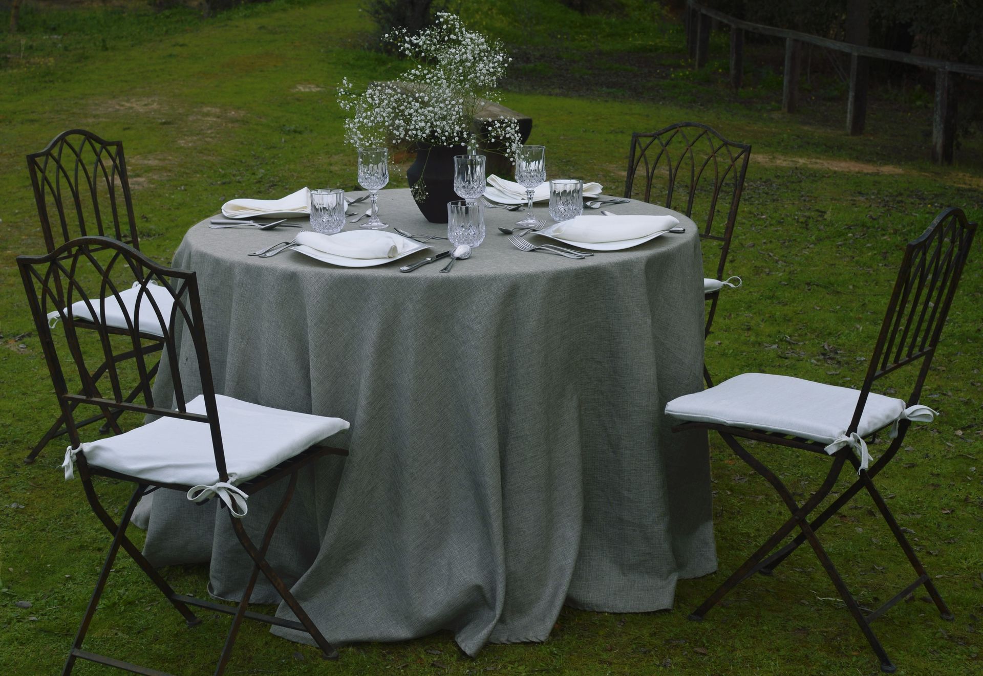 Conjunto de mesa redonda para cenar al aire libre con cuatro sillas; mantel gris y centro de mesa floral blanco.