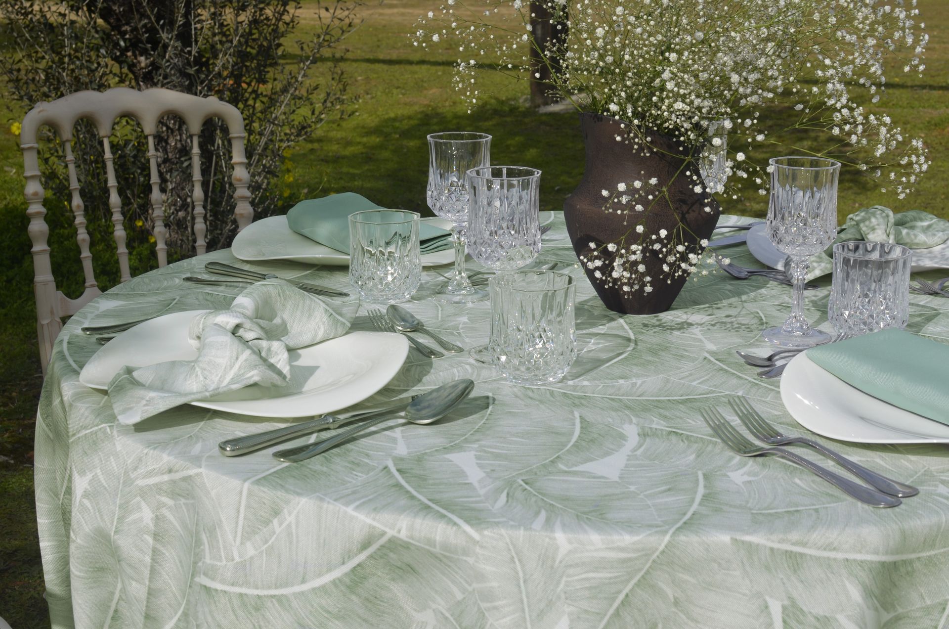 Mesa al aire libre con platos blancos, cubiertos de plata y servilletas verdes. Un arreglo floral en el centro.
