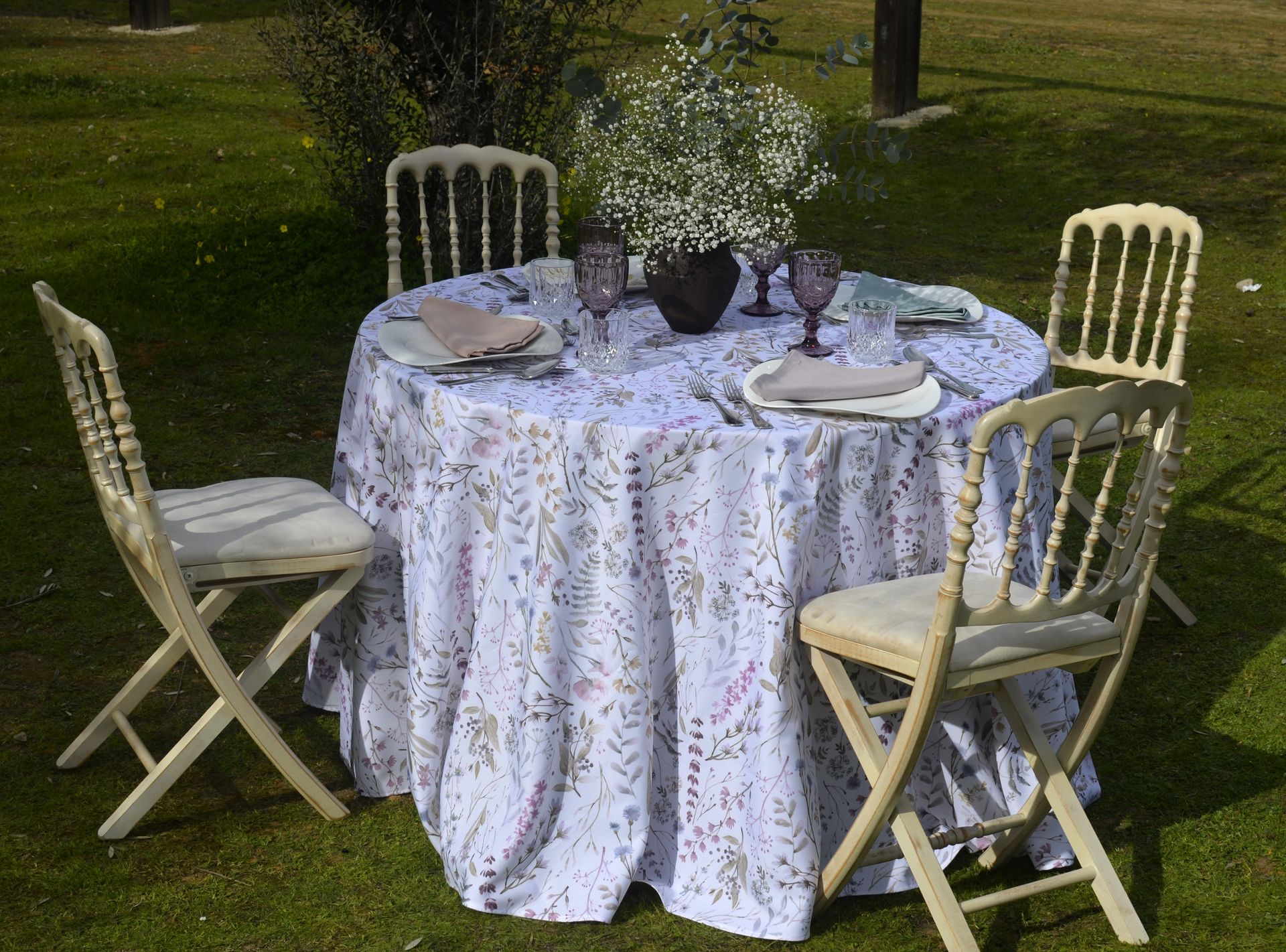 Juego de mesa redonda para cenar al aire libre; mantel floral blanco, sillas color marfil, cristalería morada y vegetación.