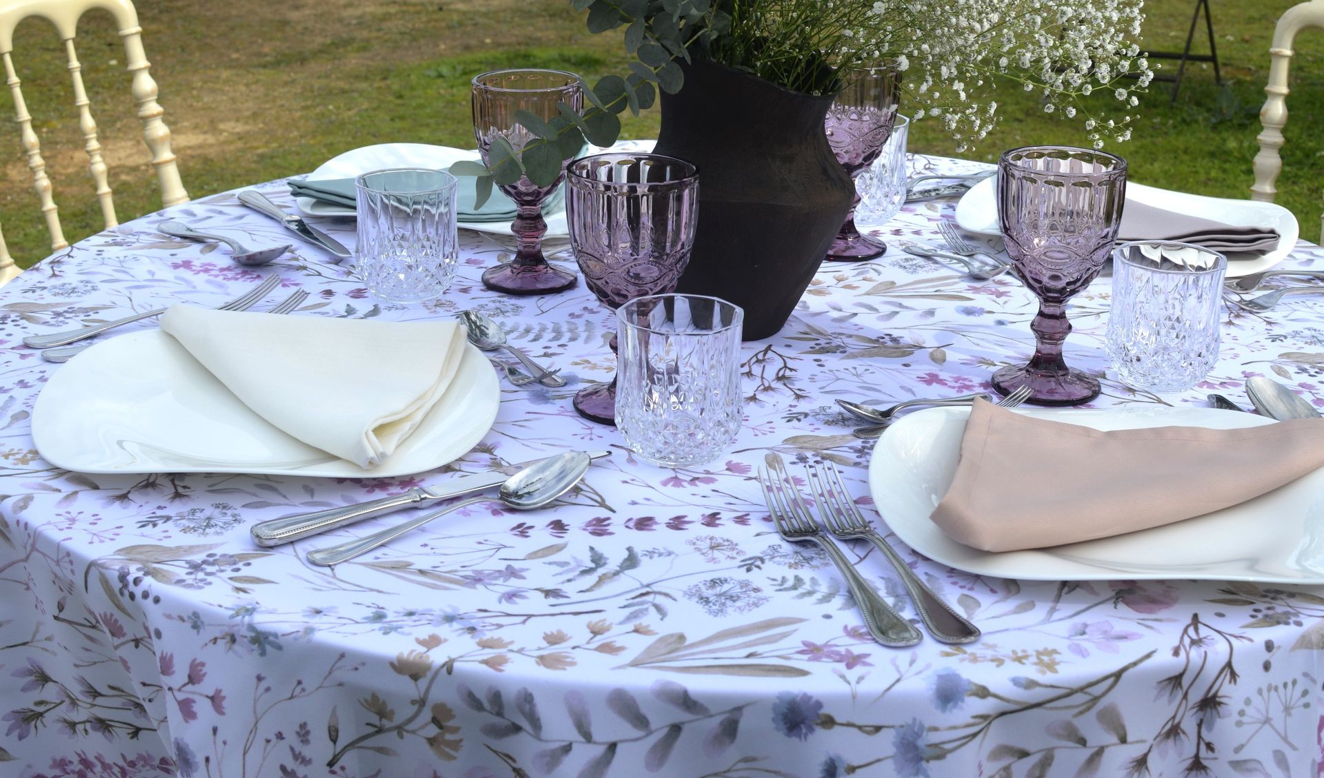 Una mesa redonda preparada para una comida al aire libre. Mantel floral, copas moradas