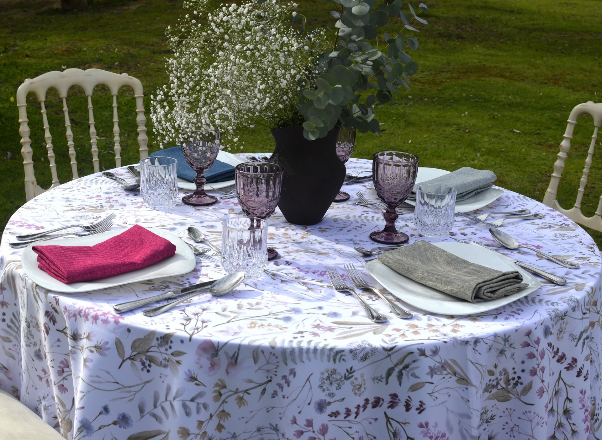 Una mesa redonda para cenar al aire libre con mantel floral, copas de vino y servilletas.
