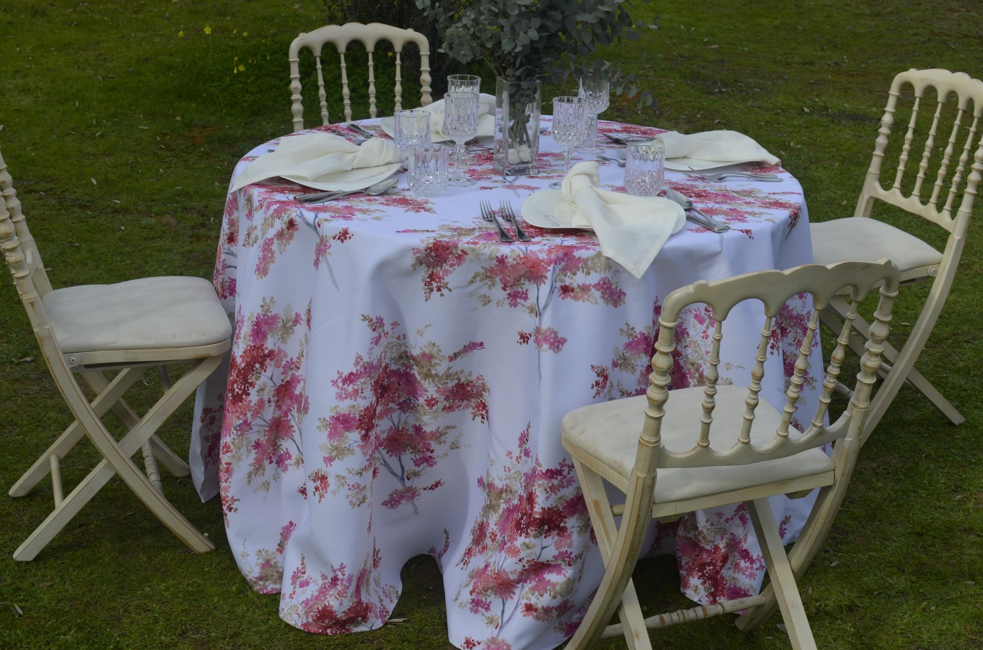 Conjunto de mesa redonda para comedor, cubierto con un mantel floral, con cuatro sillas en un entorno exterior con césped.