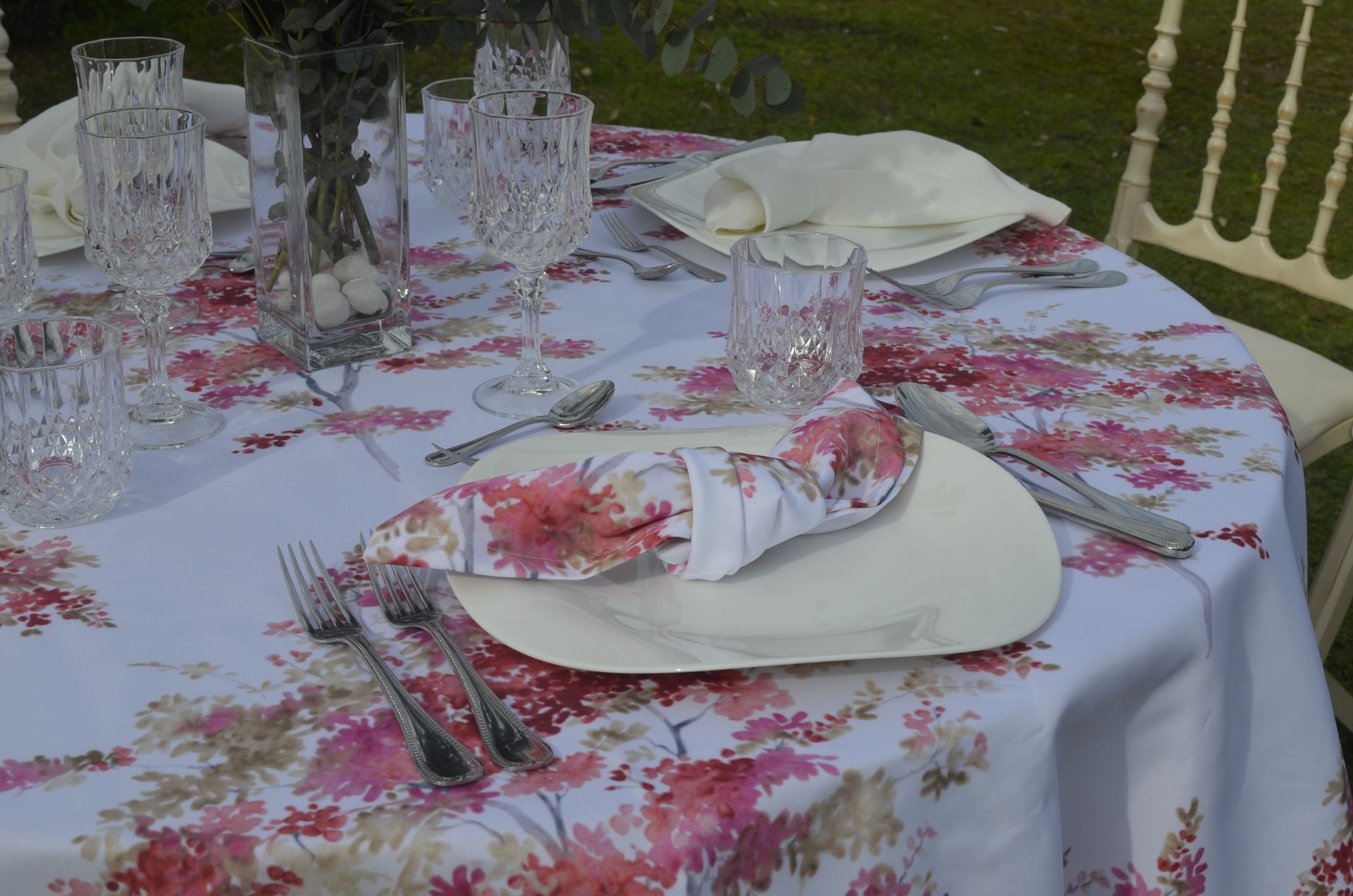 Una mesa redonda preparada para una comida formal al aire libre; mantel floral, copas de cristal y platos de marfil.