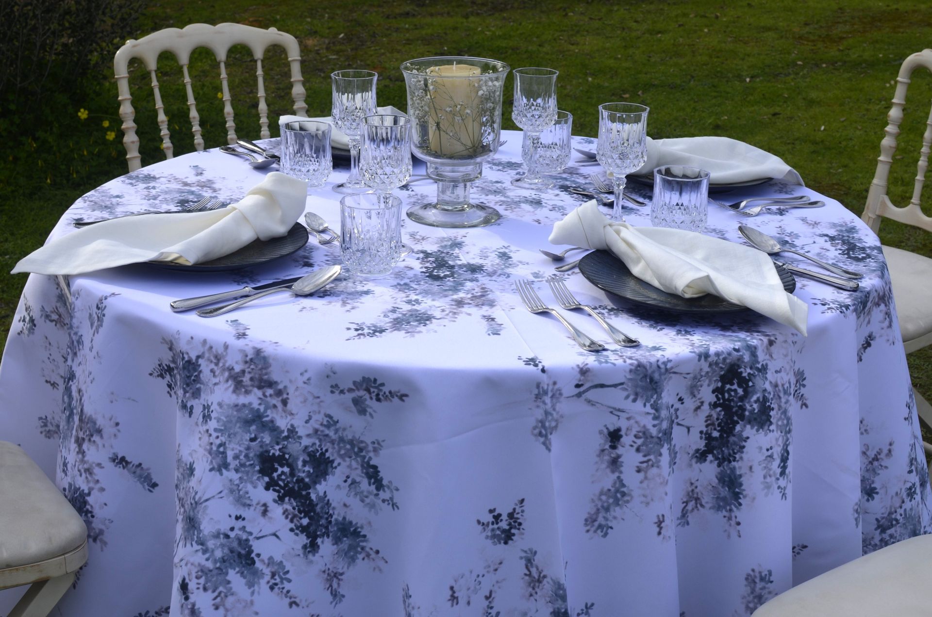 Juego de mesa redonda para cenar al aire libre con mantel floral blanco, cristalería elegante y sillas.