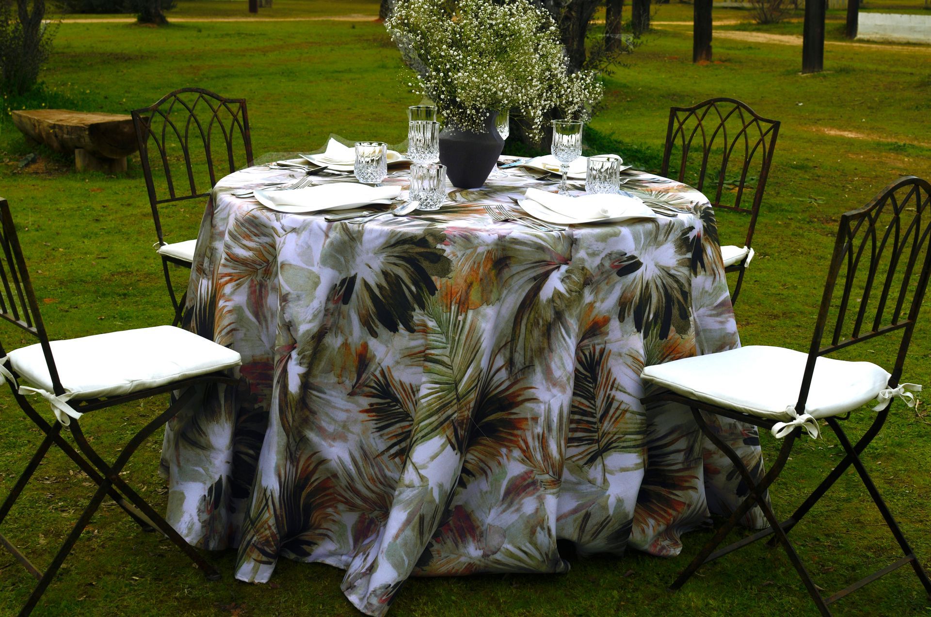 Conjunto de mesa redonda para cenar al aire libre con mantel de hojas de palma, cuatro sillas y centro de mesa floral.