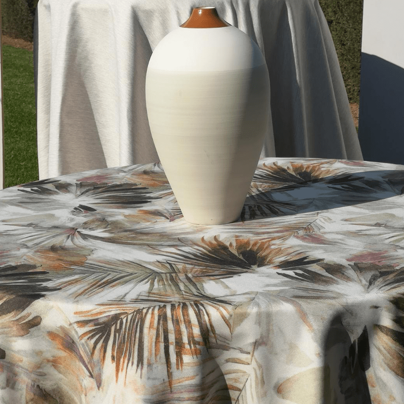 Jarrón blanco con tapa marrón sobre mantel estampado en exterior.