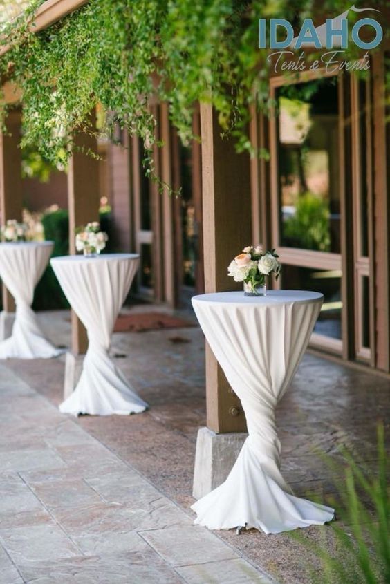 Mesas de cóctel con mantel blanco, ubicadas en el patio con centros de mesa florales. Evento al aire libre.