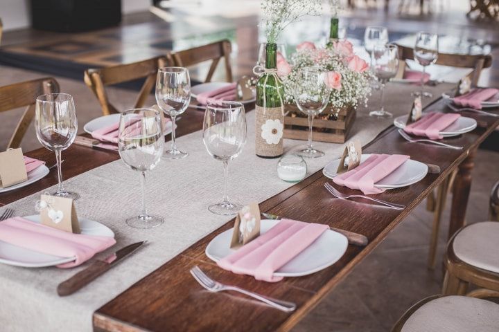 Una larga mesa de madera con platos, copas de vino y servilletas rosas.