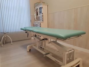 Camilla de exploración ajustable en una sala médica limpia con una superficie acolchada verde y un armario al fondo.