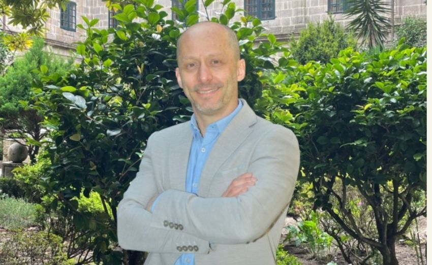 Hombre con chaqueta azul claro de pie con los brazos cruzados en un jardín frondoso.
