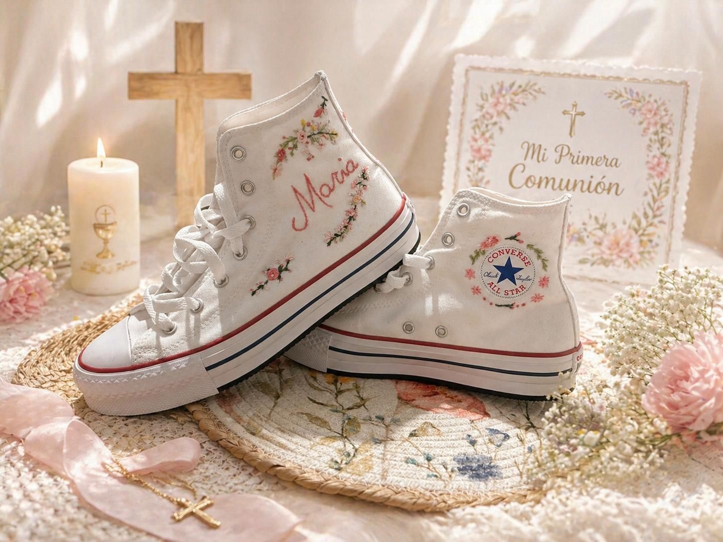 Zapatillas blancas de comunión decoradas sobre tela de encaje con una vela, una cruz y detalles florales.