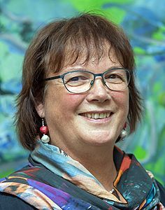 Eine Frau mit Brille und Kopftuch lächelt vor einem Gemälde.