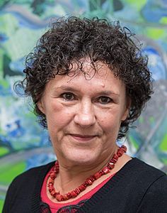 Eine Frau mit lockigem Haar trägt eine rote Halskette und ein schwarzes Hemd.