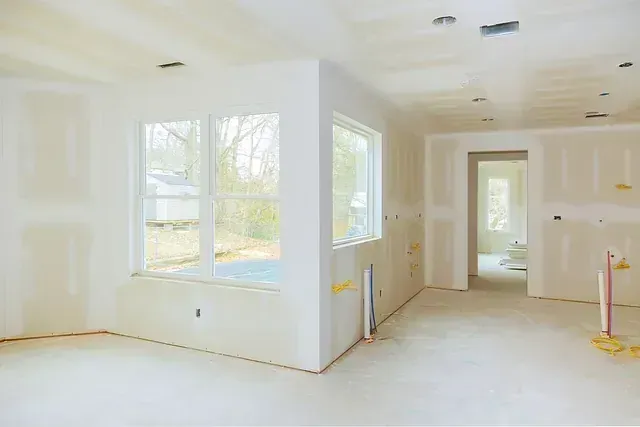 Interior de una habitación en construcción; paredes de yeso sin terminar y suelo de hormigón. Las ventanas dejan entrar la luz natural.