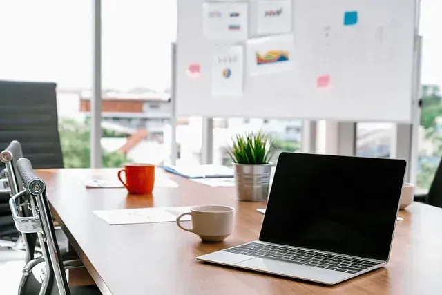 Una laptop sobre una mesa de conferencias con dos tazas y una pizarra al fondo. Habitación luminosa con vista a la ciudad.