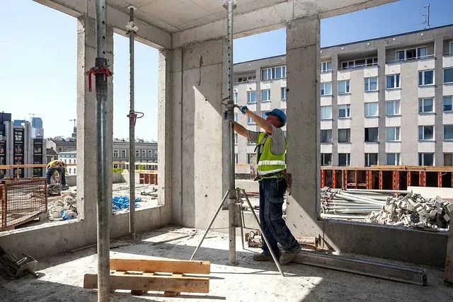 Un trabajador de la construcción con chaleco de seguridad y casco ajusta un panel en el marco de un edificio de hormigón, con vigas de soporte visibles.