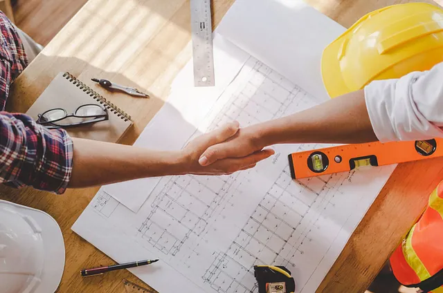 Dos personas se dan la mano sobre planos de construcción y herramientas, en señal de acuerdo o asociación.