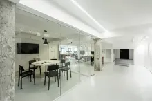 Interior de oficina moderno con paredes de cristal, sala de reuniones y un espacio de trabajo abierto visible al fondo. Paredes y suelos blancos, y una paleta de colores neutros.