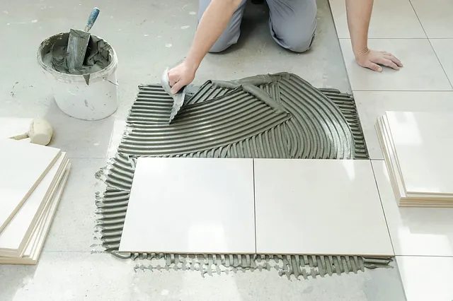 Una persona coloca baldosas en un suelo, usando una llana para extender el mortero. Se colocan baldosas blancas sobre una base de mortero gris.