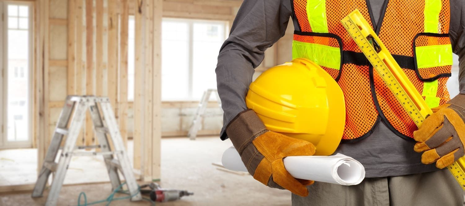 Un obrero de la construcción sostiene un casco amarillo, planos y un nivel dentro de un edificio en construcción. Lleva chaleco reflectante y guantes.