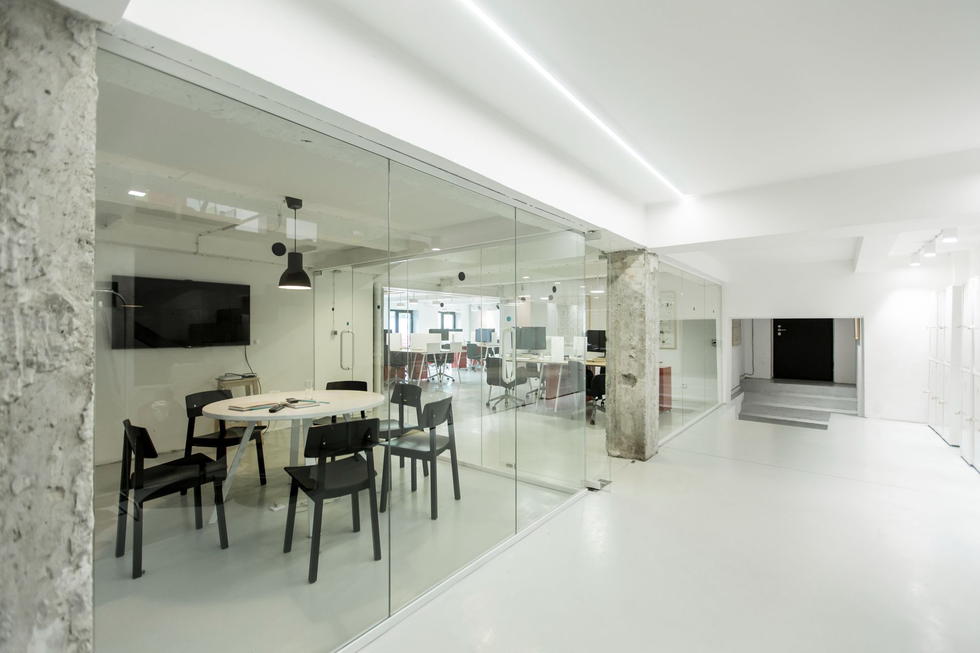 Interior de oficina moderno con sala de conferencias con paredes de vidrio y espacio de trabajo abierto con paredes blancas, pisos y pilares de hormigón a la vista.
