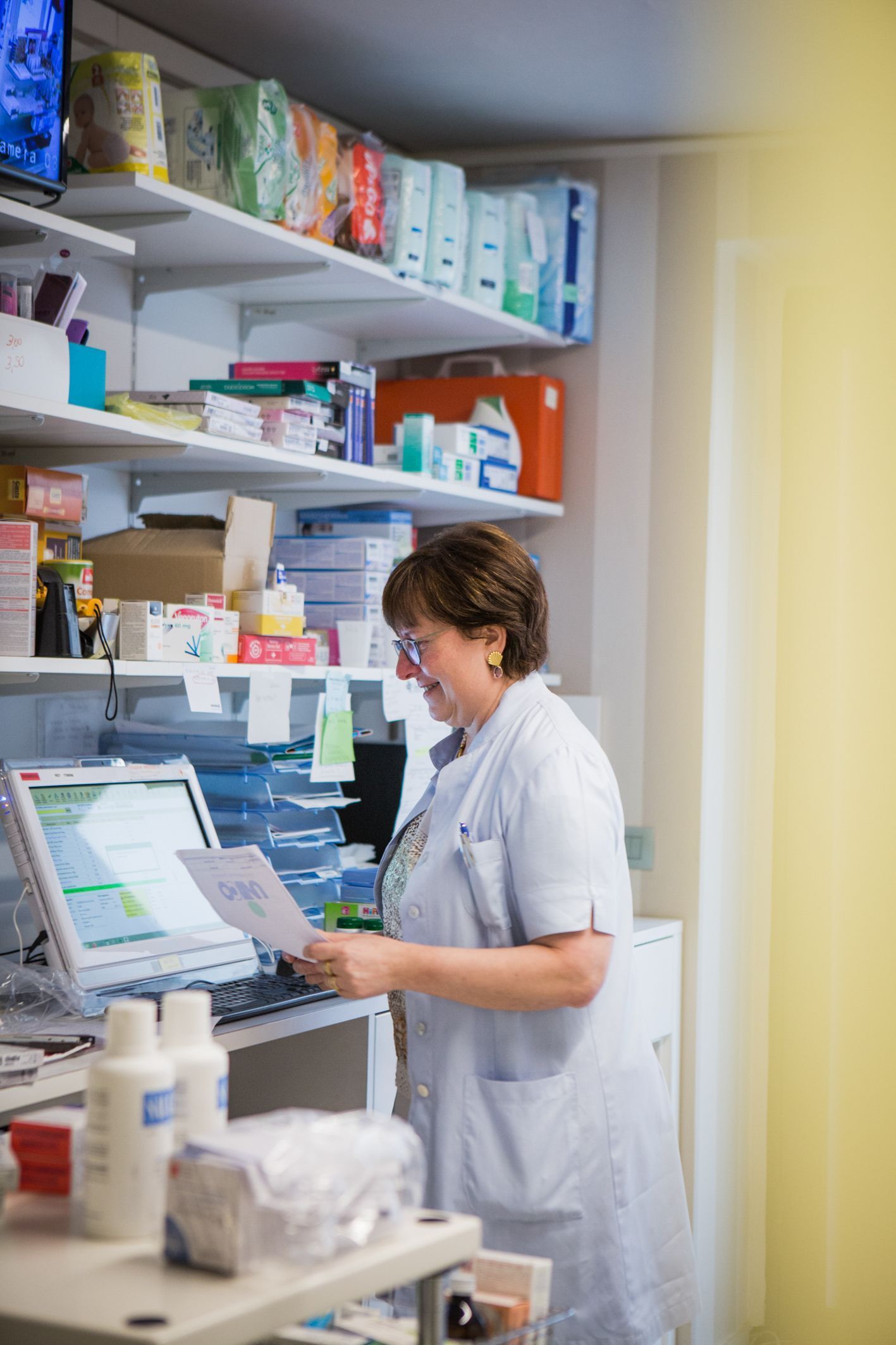 Un membro dello staff della farmacia, in camice bianco, esamina un documento mentre guarda lo schermo di un computer in un magazzino.