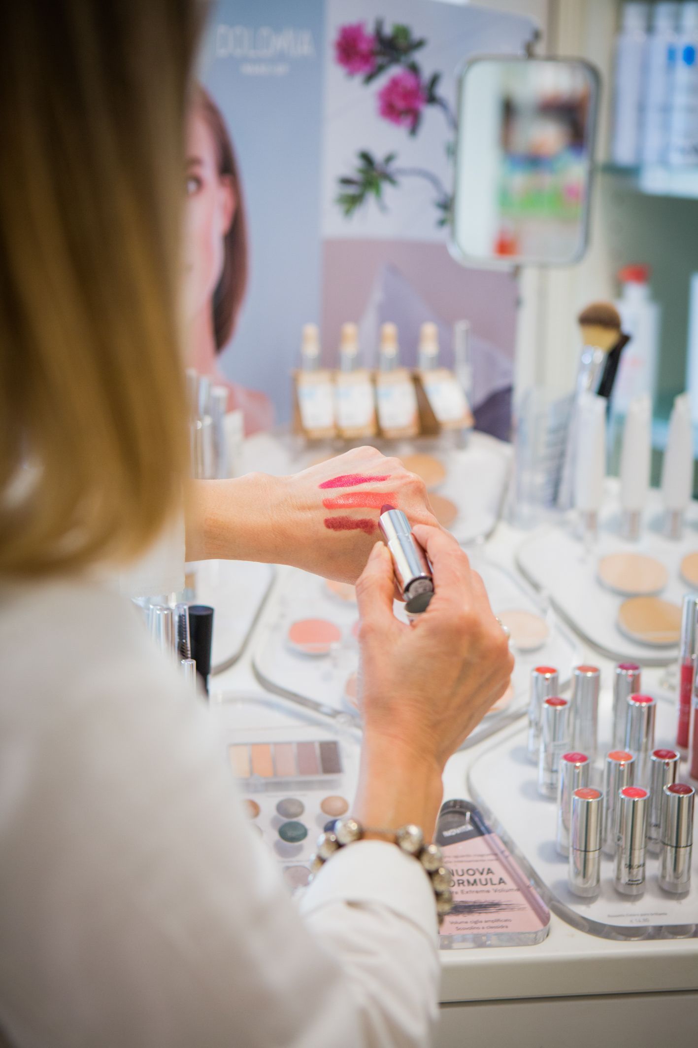 Una persona prova diverse tonalità di rossetto sulla mano davanti a un bancone espositivo di cosmetici.
