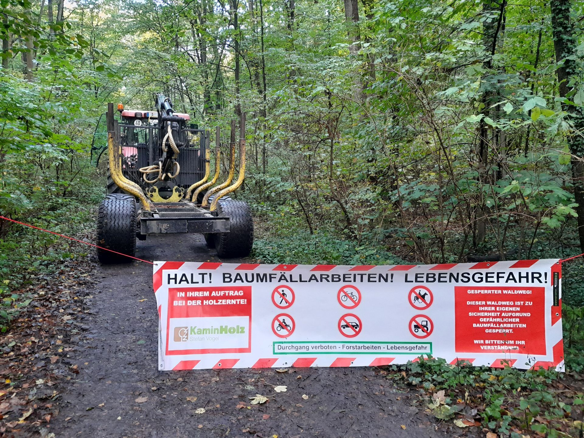 Ein Traktor fährt auf einem Feldweg neben einem Schild mit der Aufschrift „Halt! Baumfällarbeiten! Lebensgefahr!“.