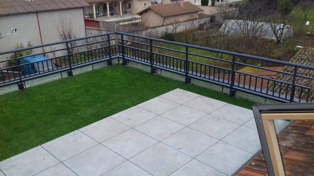 Terrasse sur le toit avec balustrades noires, pelouse verte et patio carrelé de blanc donnant sur les maisons.