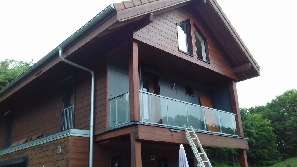 Maison en bois à deux étages avec balcon vitré et toit pentu.