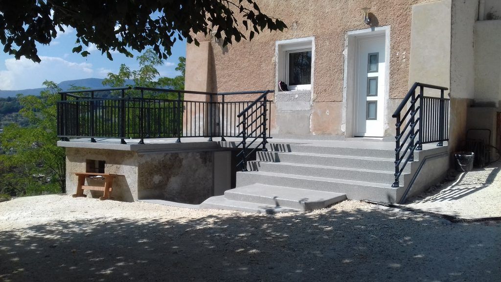 Entrée de maison moderne avec marches blanches, rambarde noire et petit balcon donnant sur une cour ensoleillée.