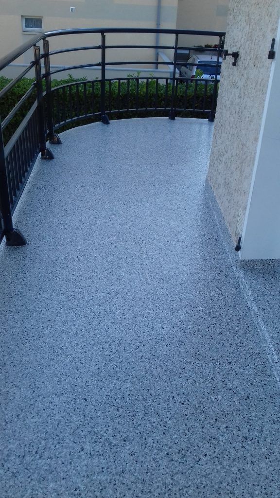 Balcon extérieur incurvé avec sol en résine, rambarde en métal noir et mur d'immeuble beige.