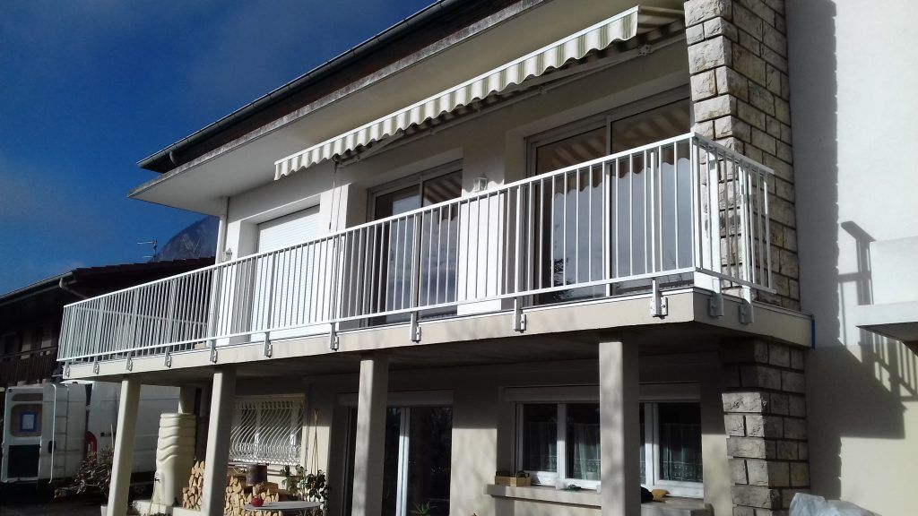 Maison à un étage avec un balcon blanc et un auvent rayé sous un ciel bleu clair.