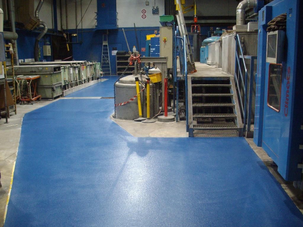 Allée en résine bleue d'un atelier d'usine avec machines, escaliers métalliques et équipements industriels.