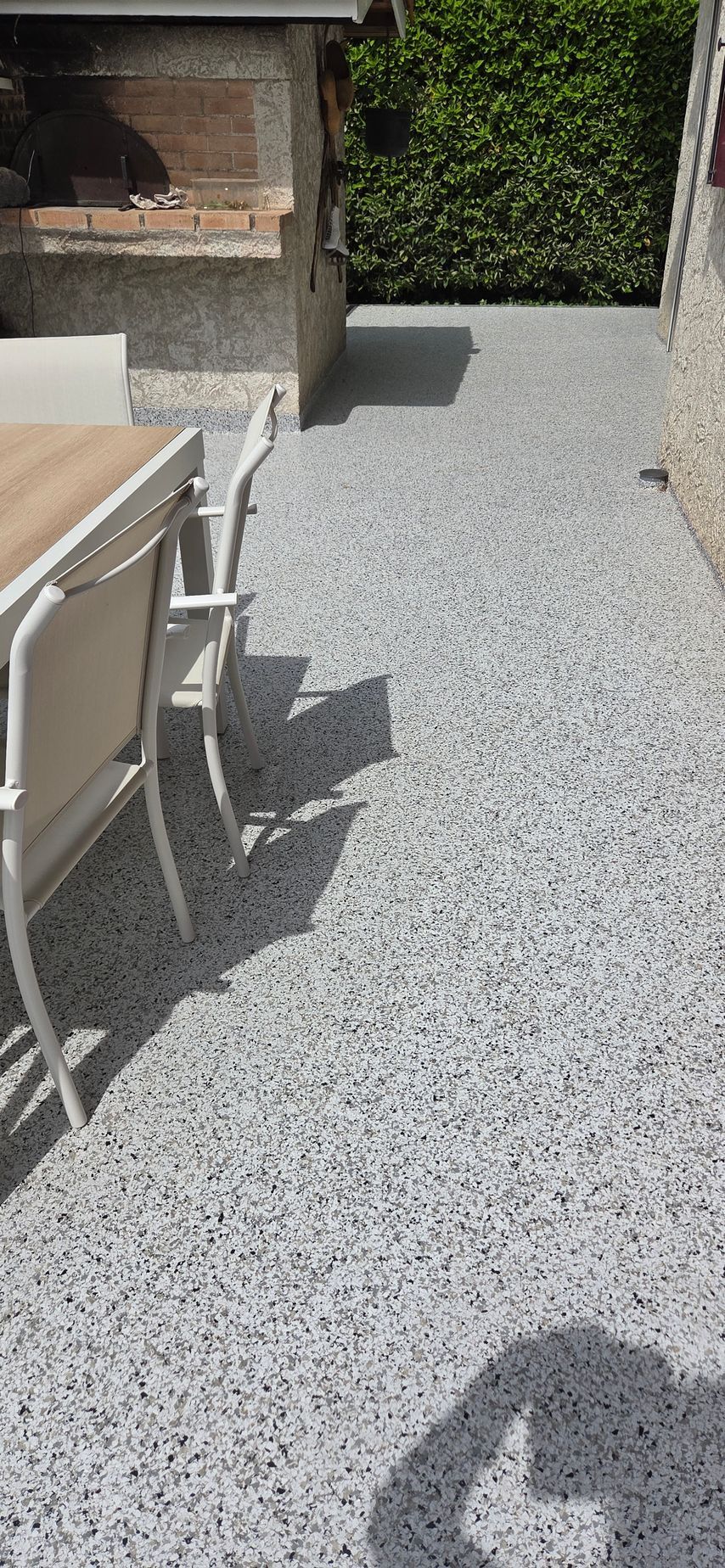 Terrasse en résine longeant un mur de briques et une haie, avec table et chaises d'extérieur au soleil.