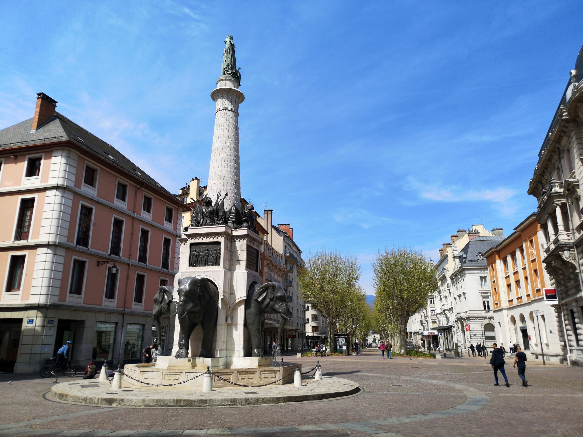 Place historique de la ville avec un haut monument en pierre, entourée de bâtiments colorés sous un ciel bleu