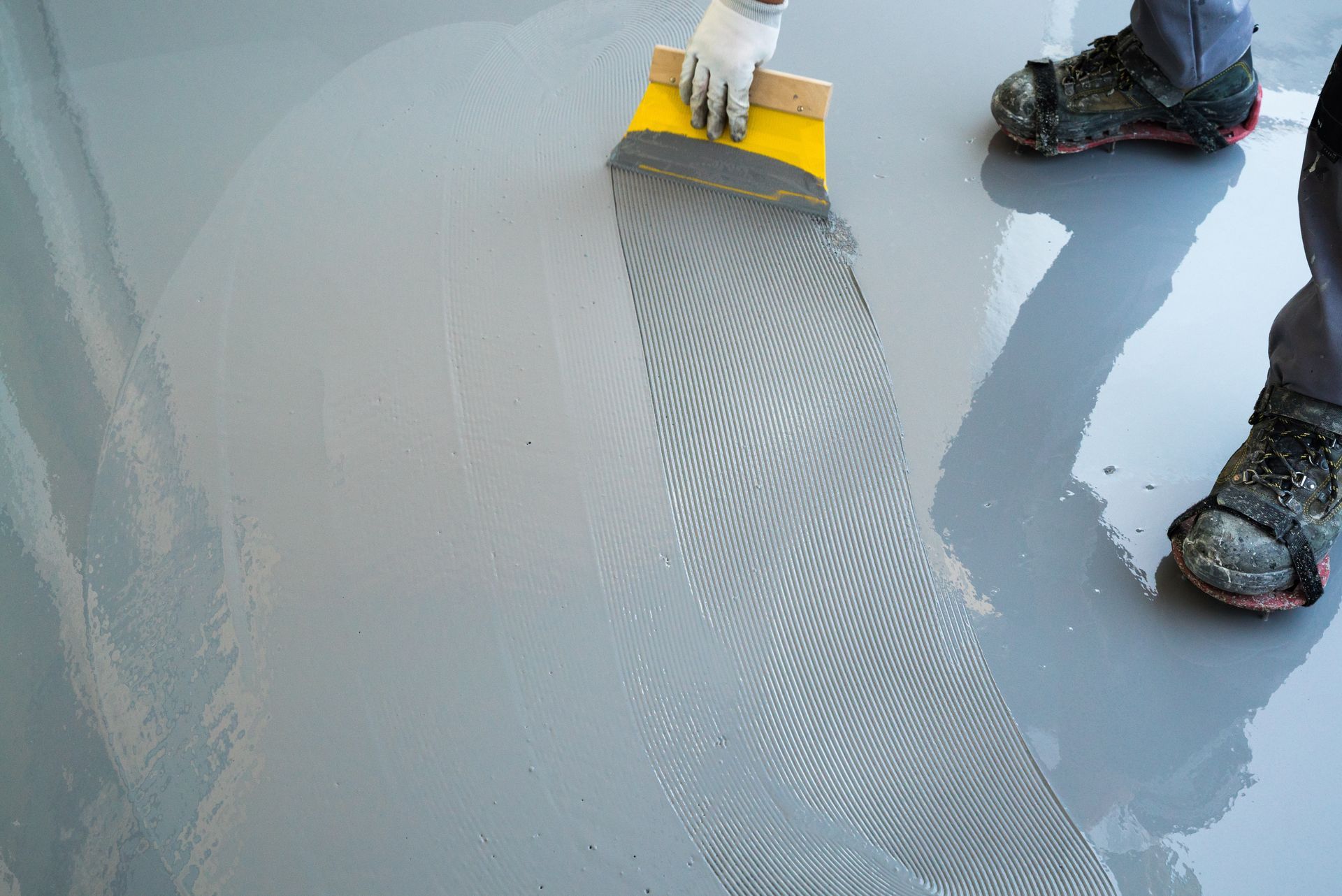 Des ouvriers étalent un revêtement liquide gris sur un sol à l'aide d'un rouleau et d'une raclette, créant ainsi une finition texturée.