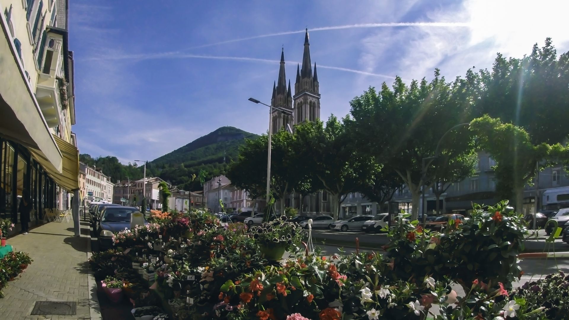 Place ensoleillée avec parterres de fleurs, arbres et une cathédrale se dressant derrière une montagne sous un ciel bleu