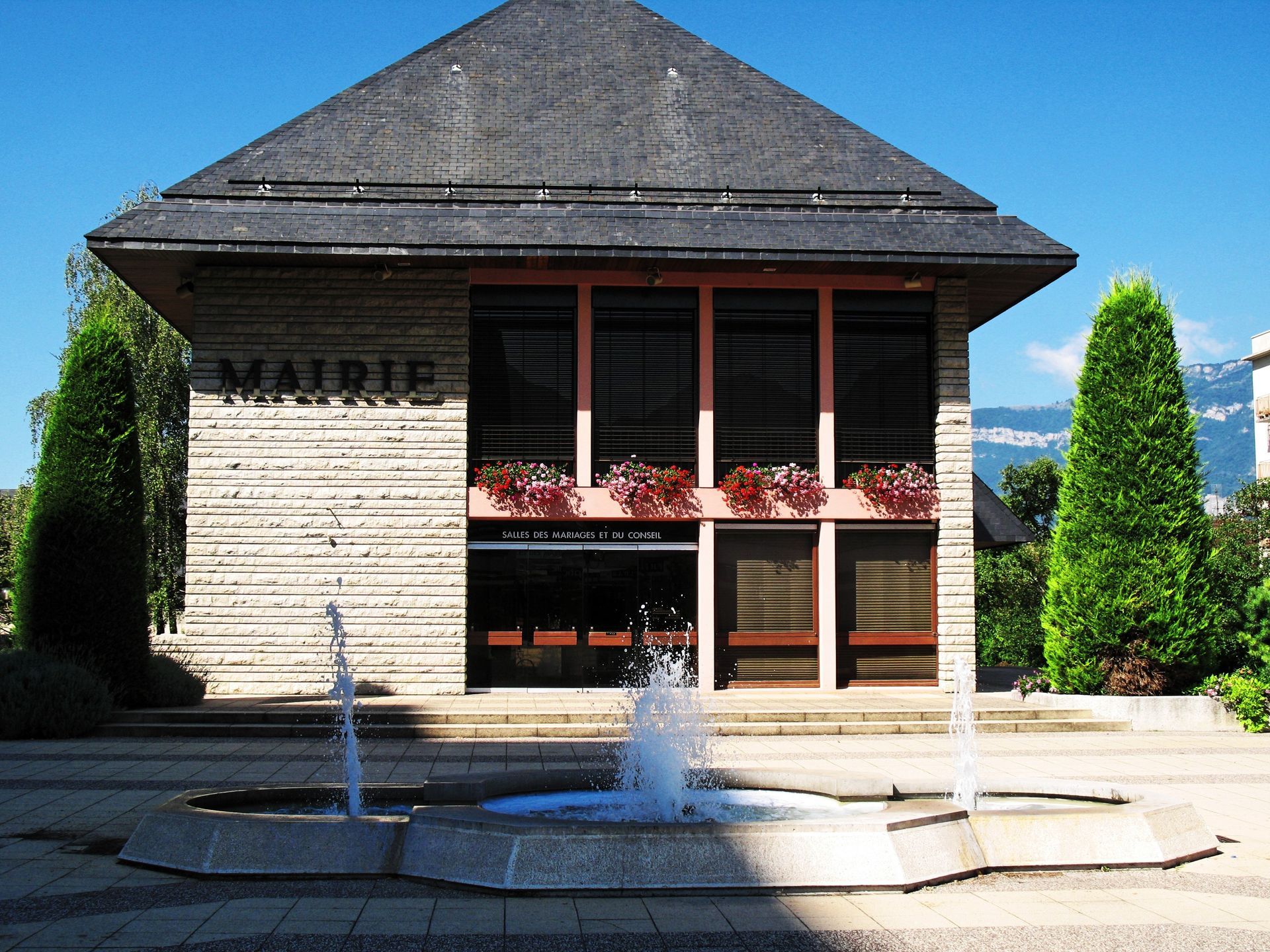 Petit bâtiment moderne avec une fontaine devant, une façade en pierre et des arbres verts de chaque côté.