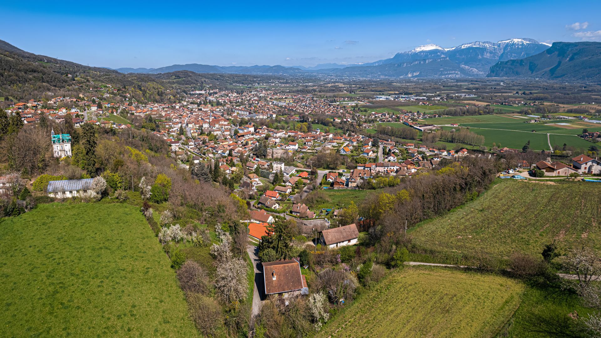 Vue panoramique d'une ville rurale avec des champs verdoyants, des collines et des montagnes enneigées sous un ciel bleu.