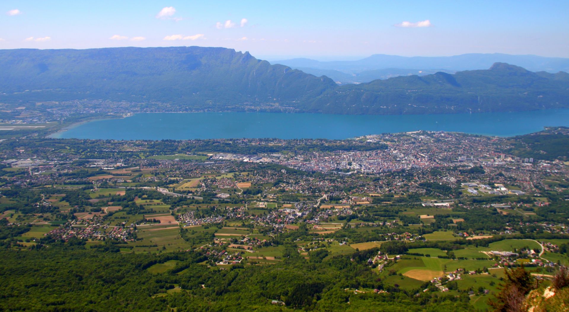 Vue panoramique d'une ville au bord d'un lac bleu, avec des champs verdoyants et des montagnes sous un ciel dégagé.