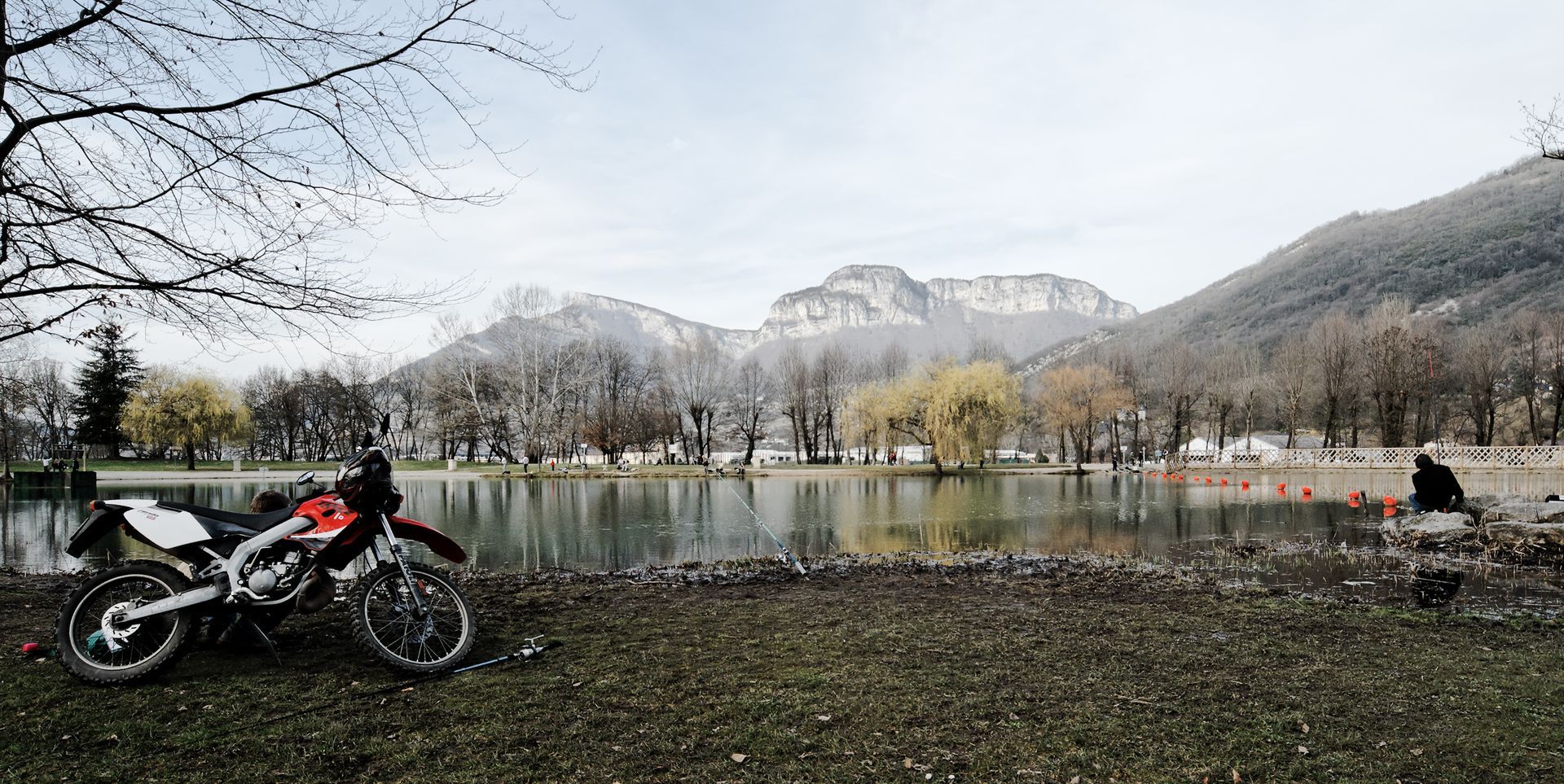 Moto garée au bord d'un lac calme, avec des montagnes et des arbres en arrière-plan.