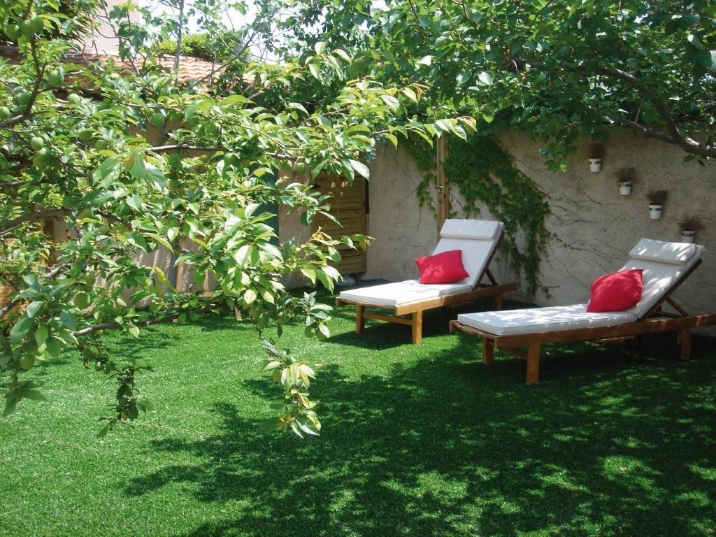 Jardin avec deux chaises longues blanches et des coussins rouges sur une pelouse ensoleillée à l'ombre des arbres.