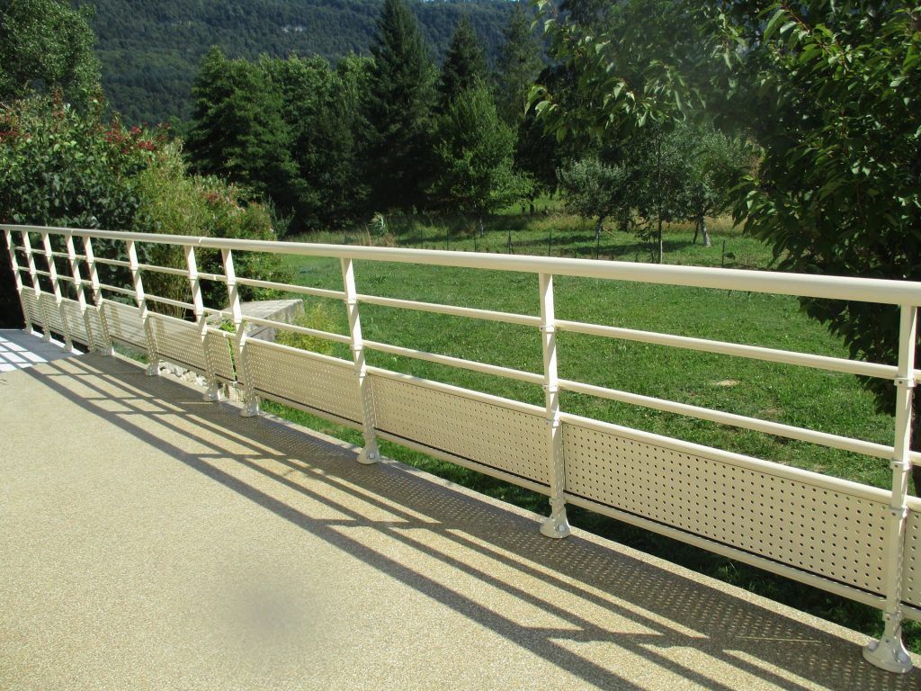 Rambarde blanche le long d'une allée ensoleillée surplombant un champ verdoyant et des collines boisées.