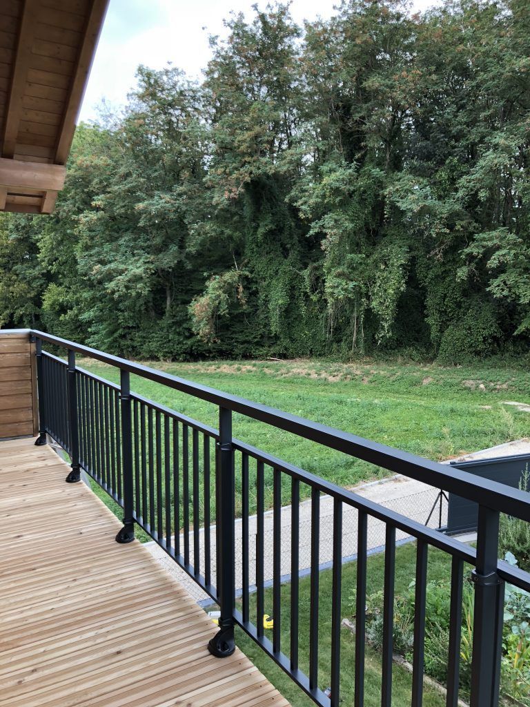 Balcon donnant sur une cour gazonnée et des arbres denses, avec une rambarde en métal noir.