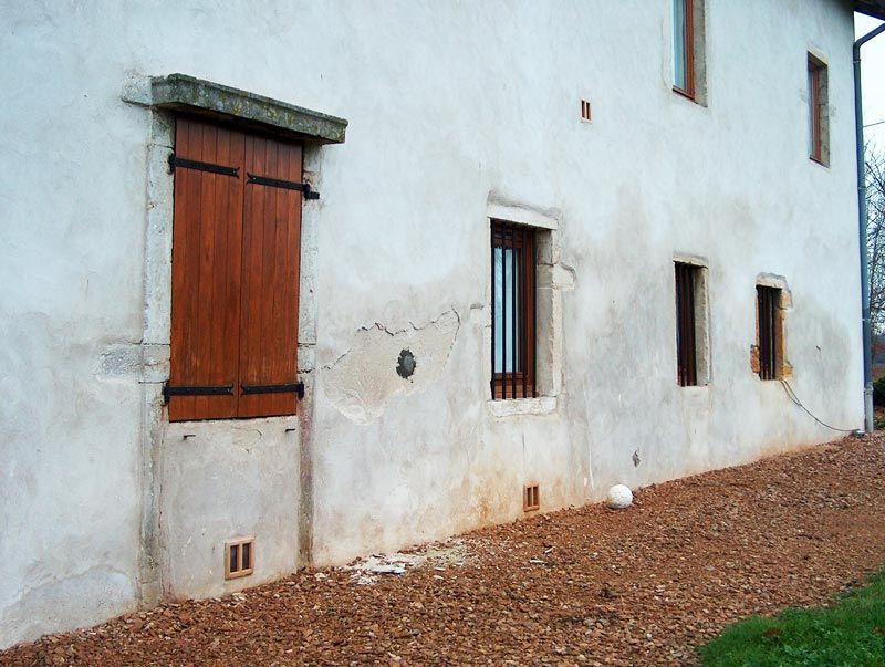 Maison à façade en stuc blanc fissuré, avec volets en bois et petites fenêtres.