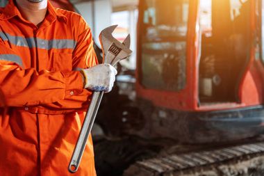 Oranssiin turvavarusteisiin pukeutunut työntekijä pitelee jakoavainta kaivinkoneen edessä rakennustyömaalla