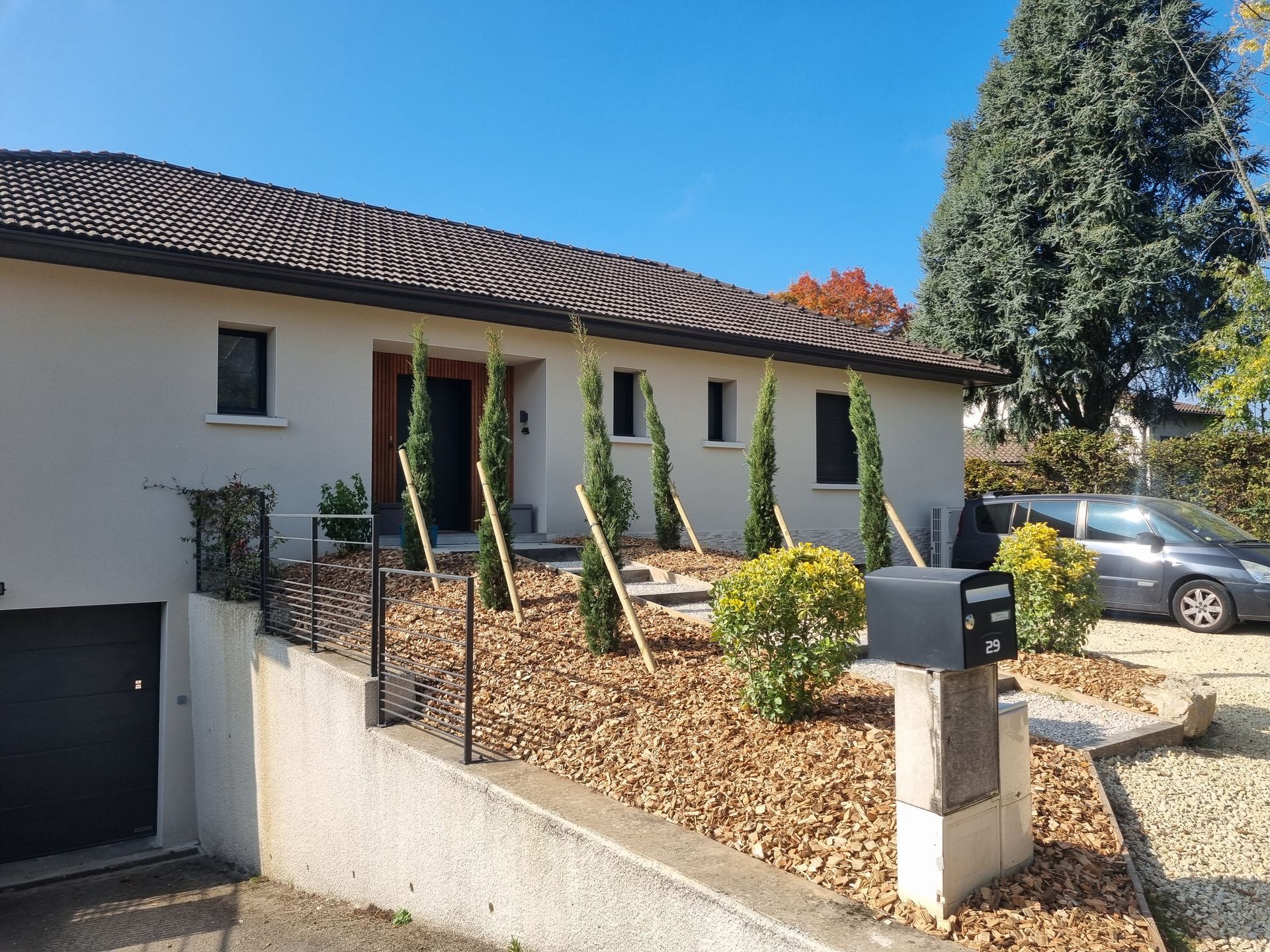 Maison blanche avec toit de tuiles, marches d'entrée, boîte aux lettres et cour gravillonnée, avec une voiture garée et un grand conifère.